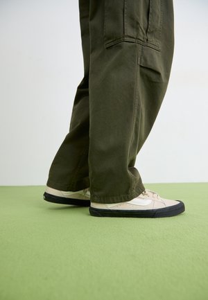 Person wearing wide-legged olive green pants and cream sneakers with black soles standing on a light green floor.
