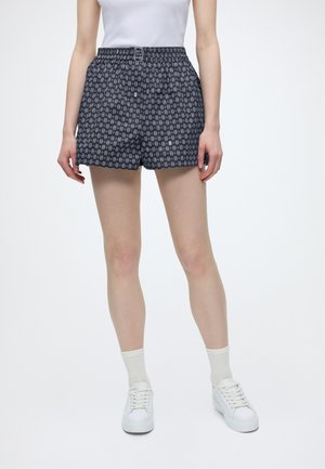 Kvinde iført navyblå mønstrede shorts med et lille bælte, hvide sokker, hvide sneakers og en hvid ærmeløs top, der står på en hvid baggrund.