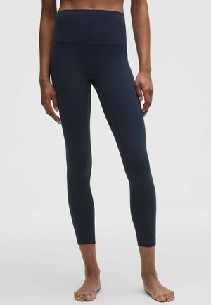 Frau trägt hoch taillierte, schwarze Leggings in voller Länge und steht barfuß vor einem schlichten hellen Hintergrund.