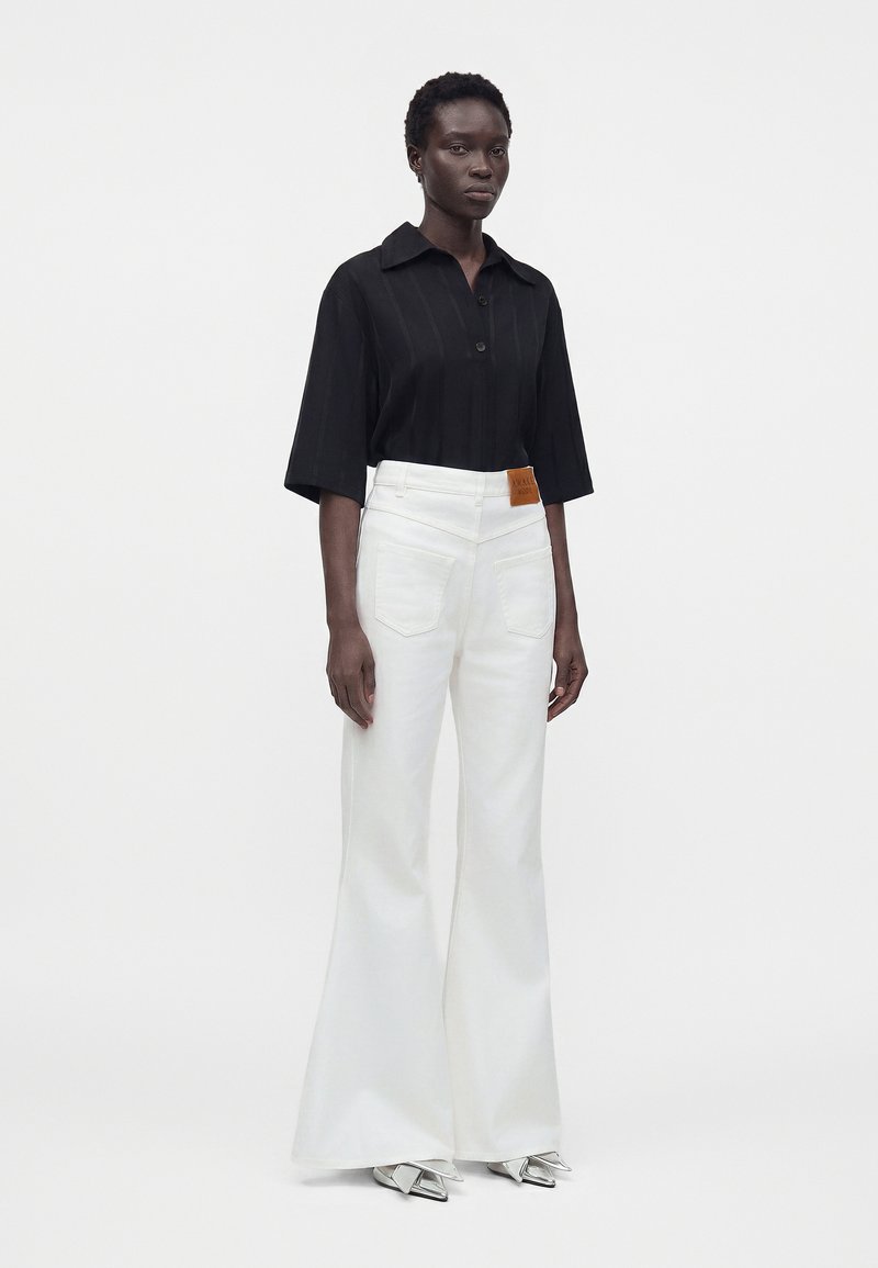 Person stands against white background wearing black short-sleeve collared shirt, high-waisted white flared pants, and silver pointed shoes.
