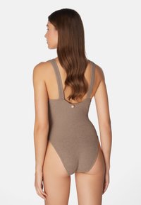 Ruskea teksturoitu yhden palan uimapuku, jossa on leveät hihnat ja nappikoriste selässä. Korkea lahkeensuu korostaa siluettia.