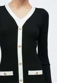 Μαύρο ριπ πλεκτό ζακέτα με λευκή επένδυση, V-λαιμόκοψη, χρυσά κουμπιά στο μπροστινό μέρος και σε δύο μπροστινές τσέπες, φορεμένη από άτομο από τους ώμους μέχρι τη μέση.