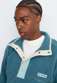 Teal fleece pullover met een crèmekleurige snap-kraag, een ritszak en lichtblauwe accenten. Beschikt over een gestructureerd oppervlak en een Columbia logo patch.