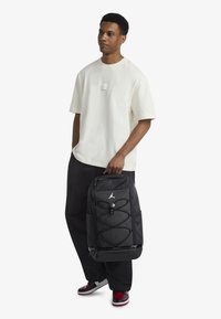 Black backpack with a smooth texture, featuring an elastic cord design, zippered compartments, and a logo. Paired with a white shirt and dark pants.