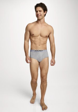 Young man with curly hair wearing gray Jockey briefs, standing barefoot on a white seamless background, looking and smiling to the side.