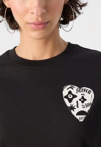 T-shirt in cotone nera con una grafica bianca a forma di cuore che presenta design facciali astratti e testo. Tessuto morbido e scollatura rotonda.