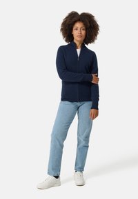 Marinefarbener Reißverschluss-Pullover mit langen Ärmeln und gerippten Bündchen, über einem weißen Hemd getragen. Kombiniert mit hellblauen Jeans und weißen Sneakers.