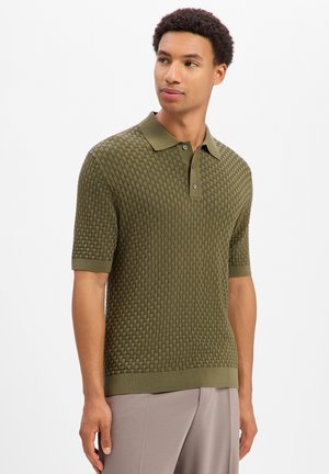 Man draagt een olijfgroen gestructureerd poloshirt met korte mouwen en lichtbeige broek, kijkt iets naar de zijkant tegen een effen achtergrond.