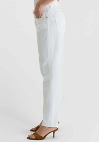 Vrouw draagt witte rechte jeans en bruine open-toe sandaal met hak, staand in profiel met hand in de zak.