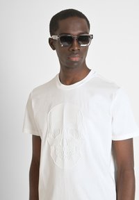 T-shirt in cotone bianco con un design di teschio in rilievo al centro. Indossato con occhiali da sole neri angolari, creando un forte contrasto.