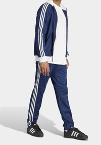 Marinblå träningsjacka och byxor med vita ränder, kombinerade med en vit t-shirt. Svarta sneakers med vita detaljer. Lättviktigt tyg, sportig design.