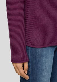 Langärmliger lila Strickpullover mit einem strukturierten Rippmuster. Der Stoff ist elastisch und weich, im Kontrast zu blauen Denim-Jeans.