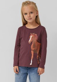 Langärmliges, kastanienbraunes Baumwollhemd mit einem grafischen Bild eines braunen Pferdes auf der Vorderseite. Rundhalsausschnitt und weicher Stoff, kombiniert mit Jeans.