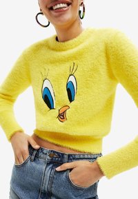 Suéter corto amarillo peludo con bordado de un personaje de dibujos animados azul y negro en el frente. Puños y dobladillo acanalados, combinado con pantalones cortos de mezclilla.