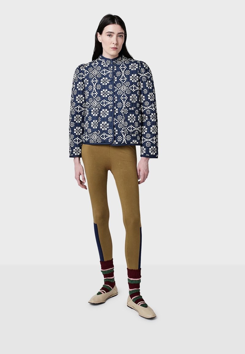 Kvinna klädd i en marinblå mönstrad quiltad jacka, senapsgula leggings med marinblå sidopaneler, randiga strumpor och beigefärgade platta skor, som står på en grå golv.