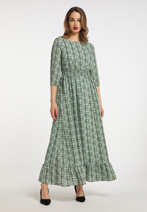 Vrouw draagt een lange groene jurk met patroon, 3/4 mouwen en een geplooide taille, gecombineerd met zwarte hoge hakken.