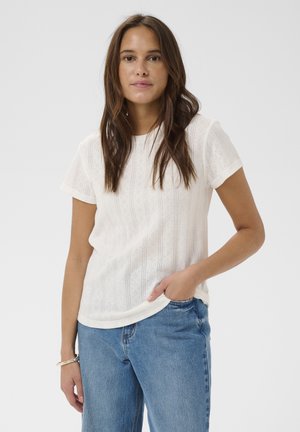 Femme aux longs cheveux bruns portant un haut blanc à manches courtes à motifs et un jean bleu, debout avec une main dans la poche, sur un fond uni.