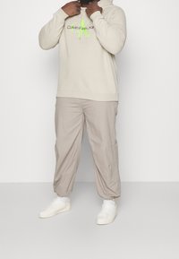 Felpa beige con logo verde neon, abbinata a pantaloni tapered beige chiaro e sneakers bianche. Il tessuto appare morbido con una vestibilità rilassata.