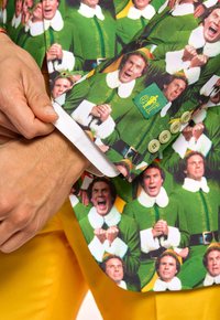 Zelený blazer s potlačou zobrazujúcou viaceré obrázky postavy v zelenom obleku. Obsahuje biely kožušinový golier a akcenty na gombíkoch.