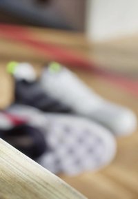 Deux chaussures de sport sont floues en arrière-plan. L'une est blanche avec des accents verts, et l'autre est noire avec des détails rouges. Un rebord en bois est net au premier plan.