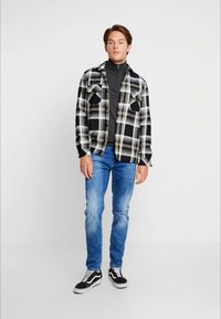 Črno-bela karo flanelasta srajca, siva zip-up jakna, modri kavbojski džins in črne superge, stoji pred enobarvno ozadjem.