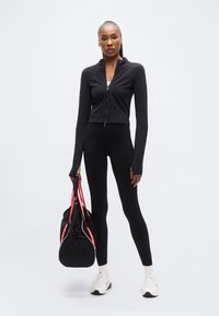 Μαύρο αθλητικό μπουφάν με φερμουάρ, μαύρο κολάν, λευκά αθλητικά παπούτσια, κρατώντας μια μαύρη τσάντα γυμναστηρίου με κόκκινες και λευκές ρίγες. Λείο, εφαρμοστό σχέδιο.