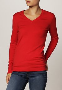 Pull à col en V rouge à manches longues avec une coupe slim, fabriqué en tissu tricot doux. Il présente des poignets et un ourlet côtelés, offrant une texture lisse.
