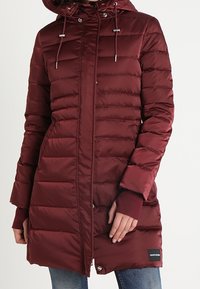 Mörkröd vadderad lång vinterjacka med huva och dragsnören, figurnära ärmar med ribbade muddarna, bärs över blå jeans.