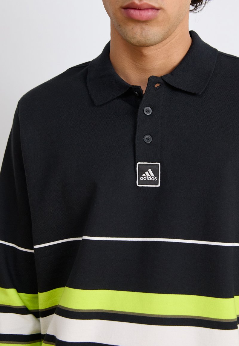 Polo noir avec des rayures blanches et vert néon, col à trois boutons, et un petit patch logo Adidas. Matériau en coton confortable.