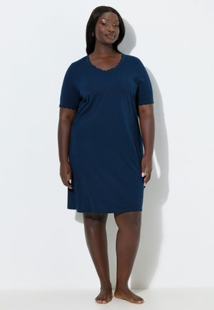 Femme debout pieds nus contre un mur blanc, portant une robe bleu marine à manches courtes arrivant aux genoux avec des bords festonnés.