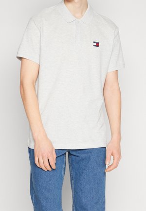 Homme portant un polo gris clair avec un petit logo Tommy Jeans et un jean bleu, debout devant un fond clair uni.