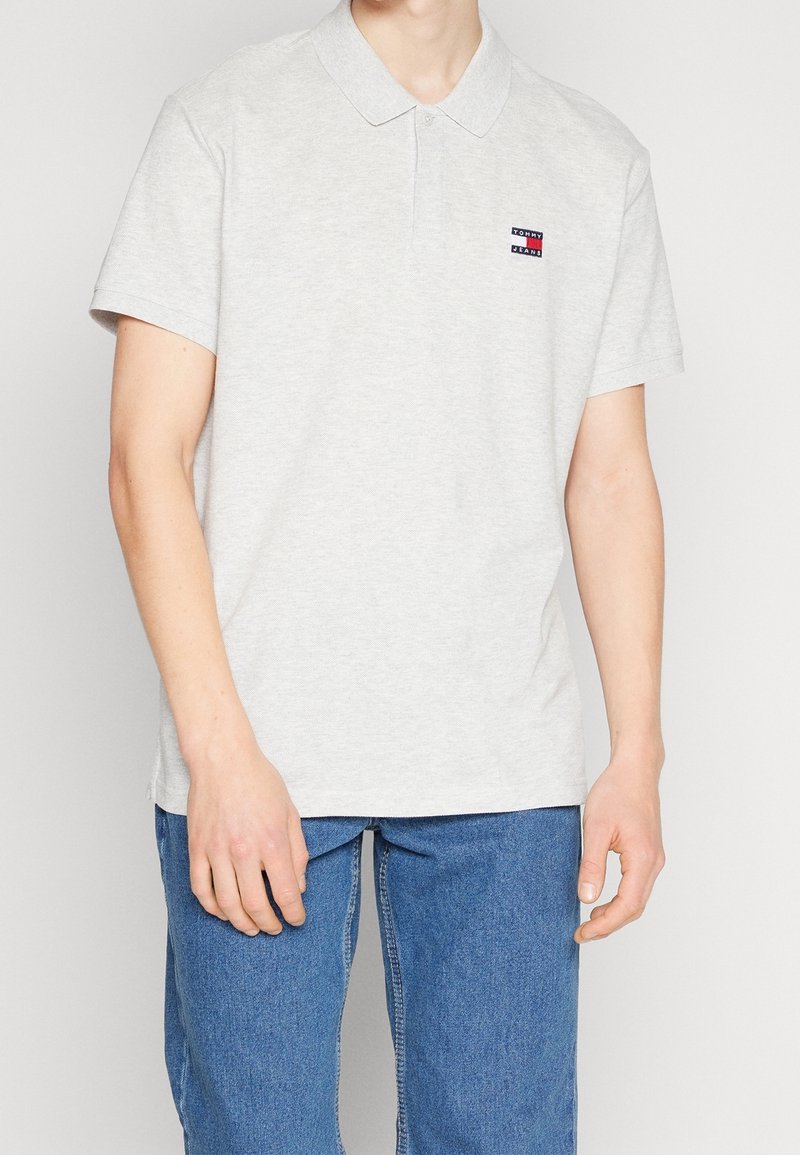 Homme portant un polo gris clair avec un petit logo Tommy Jeans et un jean bleu, debout devant un fond clair uni.