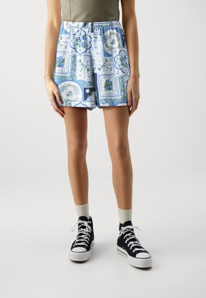 Personne portant un short bleu et blanc à motifs floraux, un haut vert olive, des chaussettes blanches et des baskets montantes noires sur un fond uni.