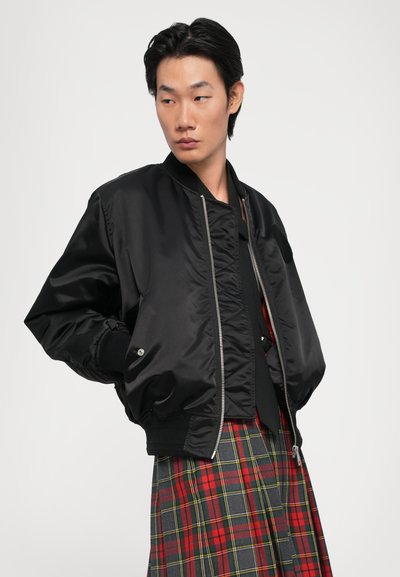 Schwarze Bomberjacke mit glänzender Oberfläche, Reißverschluss und gerippten Bündchen. Über einem rot-grauen Karorock getragen.