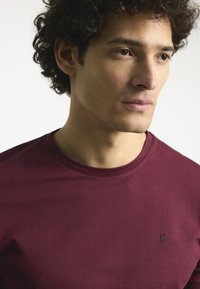 Camiseta de cuello redondo burdeos hecha de algodón suave, con un pequeño logo navy en el lado izquierdo del pecho. Mangas cortas y corte regular.