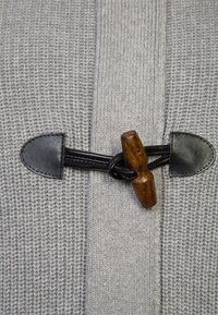 Šedá pletená látka s texturovaným vzorem, doplněná černou koženou šňůrou s dřevěným toglíkem. Akcentováno dvěma koženými konci.