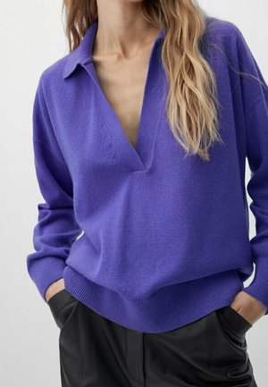 Femme portant un pull violet à manches longues avec col en V et col, pantalon noir, mains partiellement dans les poches, cheveux lâchés sur une épaule.