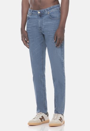 Uomo che indossa jeans azzurri a gamba dritta e sneakers bianche con dettagli marroni, in piedi davanti a uno sfondo chiaro e semplice.