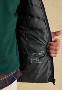 Ruka drží otvorenú čiernu prešívanú bundu, pričom je viditeľná značka Joules a symbol vodeodolnosti, zelený sveter a károvaná košeľa sú tiež viditeľné.