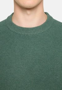 Pulover verde tricotat cu nervuri, cu gât rotund, țesătură texturată și siluetă ajustată. Apel moale cu linii orizontale constante.
