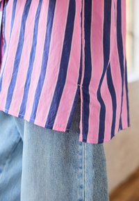 Button-up shirt with pink and navy vertical stripes, featuring a side split hem and textured fabric, worn over light blue denim.