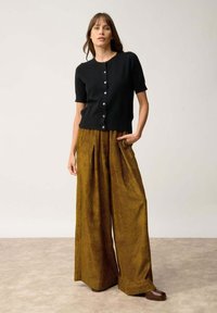 Svart kortärmad cardigan med knappar, tillsammans med vida, texturerade byxor i senapsgul kordflossa som visar vertikala ränder.