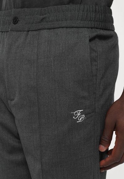Close-up of dark gray pants with elastic waistband, button, side pocket, and white embroidered "FP" initials near the pocket, worn by a person.
