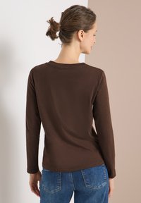 Frau mit zu einem Dutt gebundenem Haar, die ein braunes Langarmshirt und blaue Jeans trägt, steht seitlich vor einem gespaltenen weißen und beigen Hintergrund.