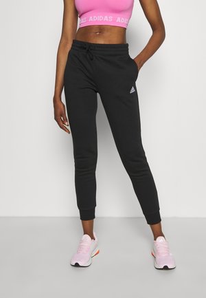 Personne portant un jogger Adidas noir, un soutien-gorge de sport rose et des baskets Adidas roses, debout devant un fond blanc uni.
