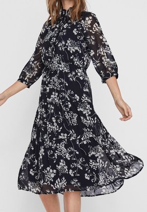 Woman wearing a black dress with white floral patterns, three-quarter sheer sleeves, and a flowing skirt.