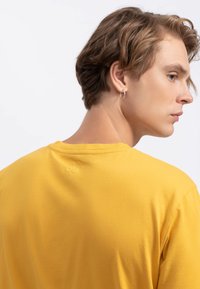T-shirt en coton jaune avec un col rond et des manches courtes. Détail notable : un petit design de cerise brodé au niveau de l'encolure au dos.