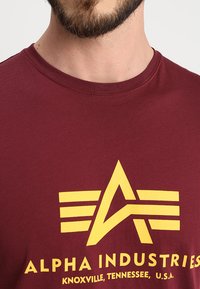 Bordeaux bomulds t-shirt med et gult grafikdesign, der inkluderer en stjerne og striber. Teksten omfatter "ALPHA INDUSTRIES" og steddetaljer.