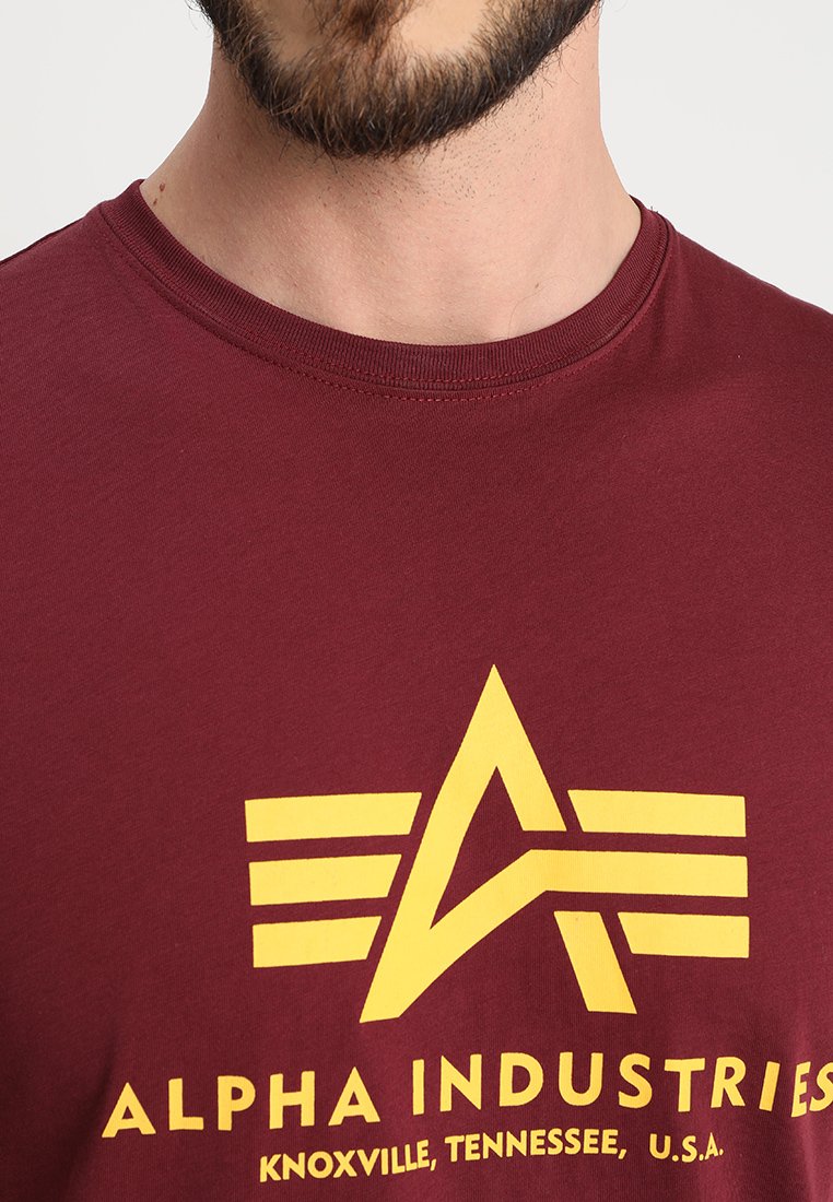 Burgundowa bawełniana koszulka z żółtą grafiką przedstawiającą gwiazdę i paski. Tekst zawiera "ALPHA INDUSTRIES" oraz szczegóły lokalizacji.