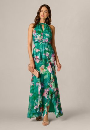 Frau in einem grünen Maxikleid mit Blumenmuster und Neckholder, die eine beige Clutch hält und mit einem Fuß leicht vorangestellt vor einem einfarbigen Hintergrund steht.